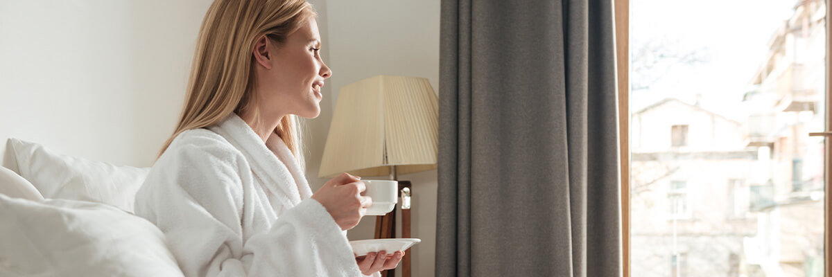 Woman sitting on bed in a robe, holding a coffee and looking out window.